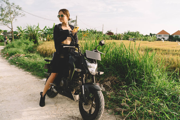 Full body of positive young slender woman in black clothing and sunglasses using mobile phone and looking away while sitting on bike and resting - Fotó, kép