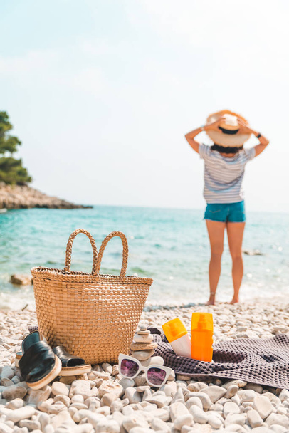 пляжний предмет солом'яний капелюх і сумка з лапками і сонцезахисним кремом на березі моря жінка на фоні
 - Фото, зображення