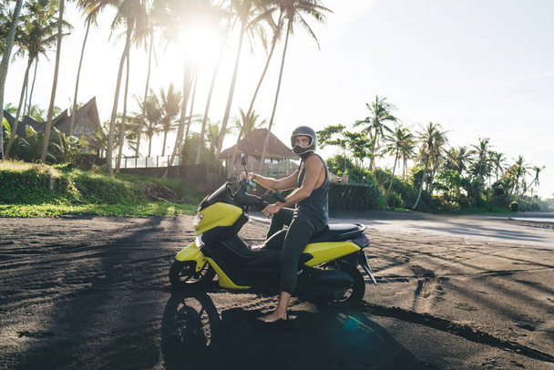 Full length of anonymous male biker wearing safety helmet and casual clothes sitting on scooter near high palms growing on sandy beach - Photo, image