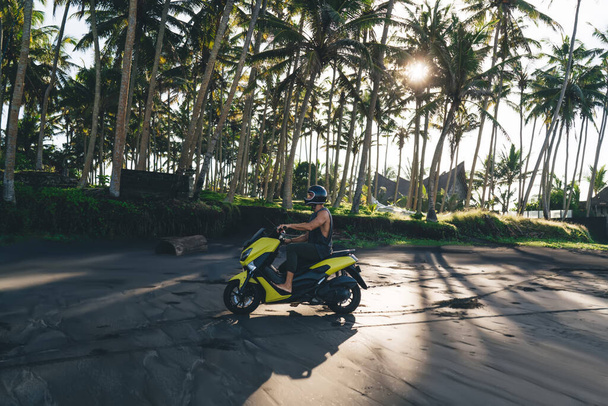 Full length of anonymous male biker wearing helmet and casual outfit driving motorbike along palm trees on sandy beach during sunset - Valokuva, kuva