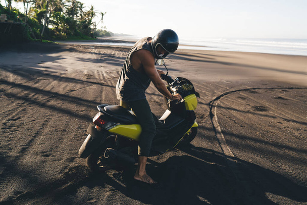 Side view full body sporty male biker in helmet standing with modern motorcycle on sandy seacoast in sunny exotic country - Fotoğraf, Görsel