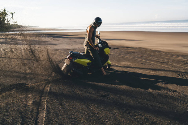 Longitud completa de motocicleta masculina irreconocible que monta la motocicleta en la playa arenosa cerca del mar tranquilo en el día soleado del verano cerca de los árboles tropicales - Foto, imagen