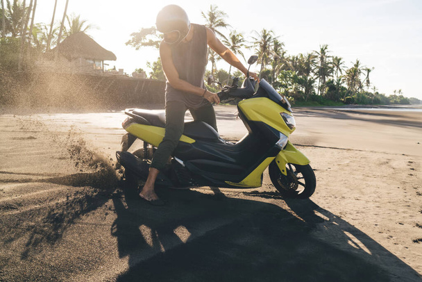 Full length of unrecognizable male biker wearing protective helmet and casual clothes getting stuck in sand while driving along seashore - Foto, afbeelding