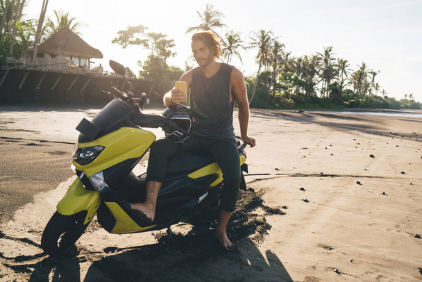 Full length of bearded man sitting on motorcycle on sandy seashore near tropical resort while surfing mobile phone on sunny day - Фото, изображение