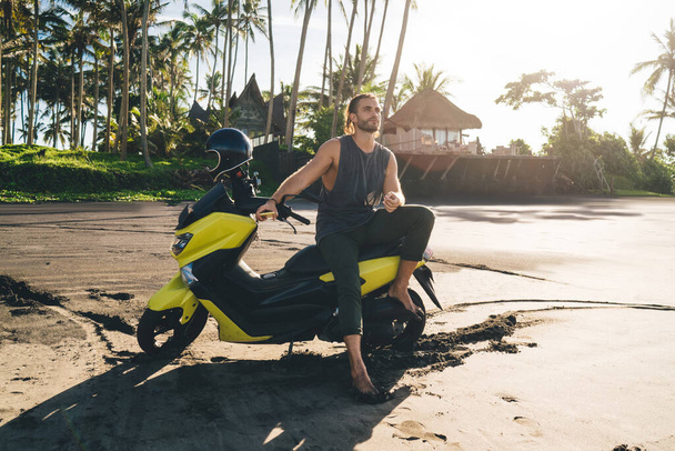 Full body of man with phone sitting on motorbike among sandy beach and enjoying summer holidays while spending time in tropical country and looking away - Foto, Imagen