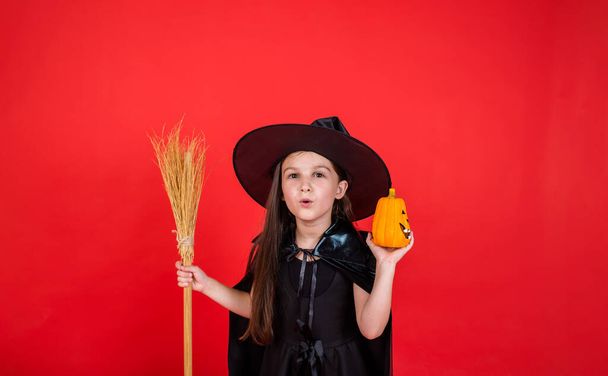 ένα μικρό μελαχρινό κορίτσι σε ένα κοστούμι μάγισσα με ένα καπέλο κρατά μια σκούπα και μια κολοκύθα σε ένα κόκκινο φόντο με ένα αντίγραφο του χώρου - Φωτογραφία, εικόνα
