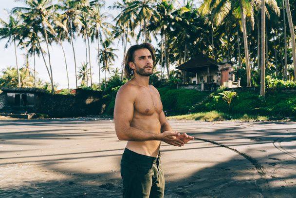 Calm muscular bearded guy with naked torso relaxing on sandy tropical beach with palms and guest house and rubbing hands in summer hot day - Φωτογραφία, εικόνα