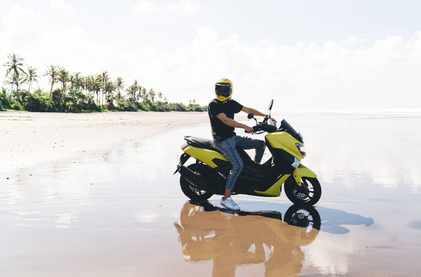 Повне тіло анонімного чоловіка-мандрівника в шоломі, що сидить на мотоциклі на мокрій піщаному пляжі, проводячи час наодинці в тропічному курорті
 - Фото, зображення