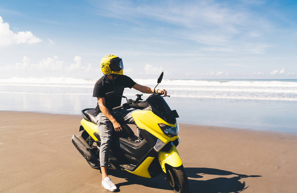 Faceless male in casual clothes and helmet riding motorbike against scenic seascape and cloudy blue sky on sunny summer day - Фото, изображение