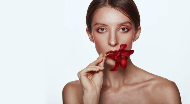 Pretty woman holding red flower in mouth naked shoulders clear skin model with pink makeup. Skin care and Spa concept on white background. Woman looking at camera, space for text - Photo, Image