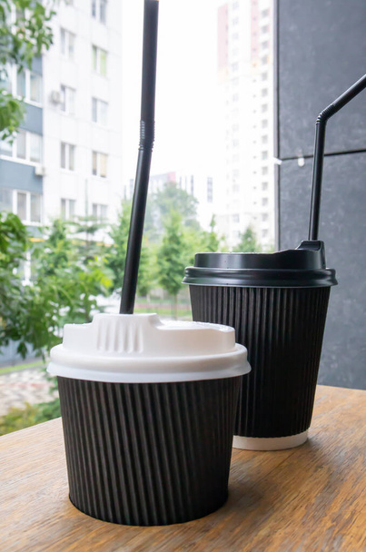 Дві чорні паперові чашки з кришками на відкритому повітрі влітку в сонячну погоду на дерев'яному столі кафе, кав'ярні або ресторані
 - Фото, зображення