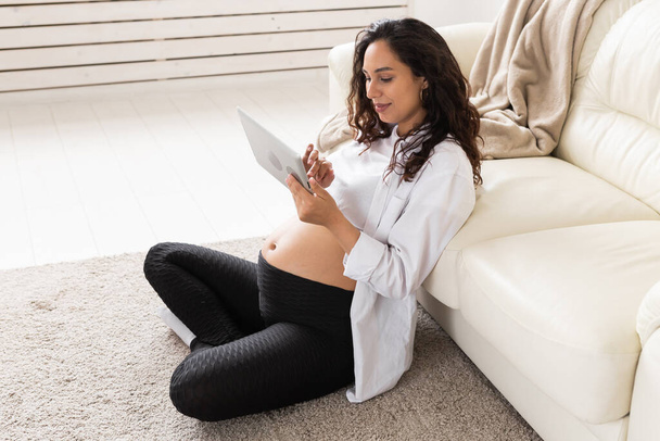 Schwangere Frau mit Tablet-Computer sitzt in der Nähe von Sofa zu Hause. Schwangerschaft und Informationen zum Elternschaftskonzept. - Foto, Bild