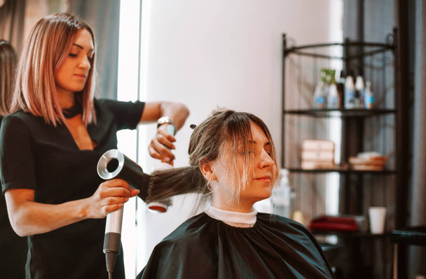 Happy woman with stylist making hairdo at salon. Competent female hairdresser deciding what to do in hair salon with brunette female client. Two beautiful ladies discussing new hairstyle. Professional coiffure and stylist work concept - Photo, Image