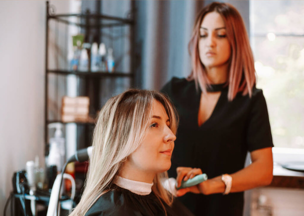 Szczęśliwa kobieta ze stylistką robiącą fryzurę w salonie. Kompetentna fryzjerka decyduje, co robić w salonie fryzjerskim z brunetką klientką. Dwie piękne panie rozmawiające o nowej fryzurze. Profesjonalna koncepcja coiffure i stylista pracy - Zdjęcie, obraz