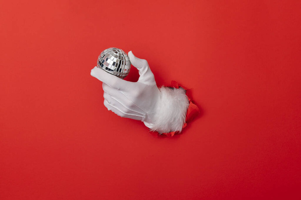 Santa Claus hand with disco glitter ball through a hole in red paper background - Foto, Imagem