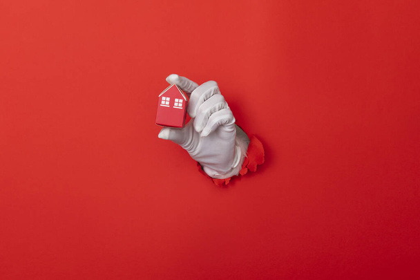 Santa Claus hand holding model house through a hole in red paper background - Photo, image