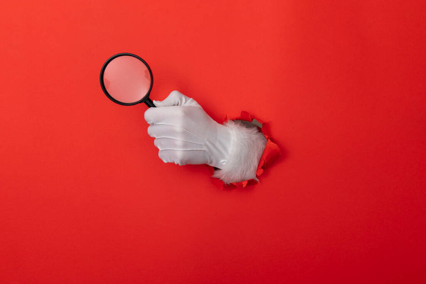 Santa Claus hand with magnifying glass through a hole in red paper background - 写真・画像
