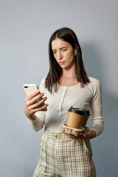 A woman in a white sweatshirt holds two cups of coffee in one hand, and a smartphone in the other. Tea or coffee to take away. Remote communication - Photo, Image