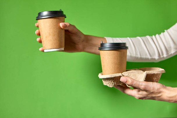 Hands holding two cups brown paper with black lid. Two coffee special offer or promo. Hands holding two cups on green background. Tea or Coffee to go. Brown paper cup with black lid. - Photo, Image