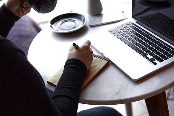 Egy fiatal lány ül egy kávézóban, egy laptopon dolgozik és jegyzetel egy jegyzetfüzetben. Close - a kezek fényképe. A szabadúszó és távoli munkavégzés vagy képzés fogalma. - Fotó, kép