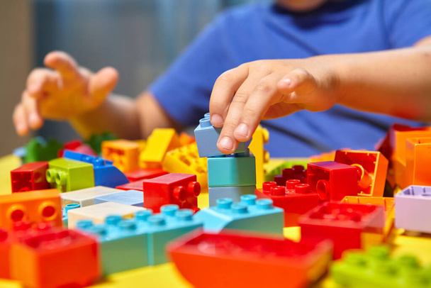 A beautiful boy is playing at home with building blocks. A cute smiling boy is playing with a constructor with a lot of colorful plastic blocks in the room, building a city. Preschool classes. - Photo, Image