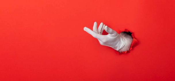 Santa Claus hand wearing white glove through a hole in red paper background - Φωτογραφία, εικόνα
