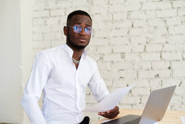 Hombre africano sonriente que trabaja en una oficina moderna detrás de un portátil y mira a la cámara. Hombre de negocios afroamericano guapo en camisa y pantalones de vestir utiliza el ordenador portátil y toma notas mientras trabaja en - Foto, Imagen
