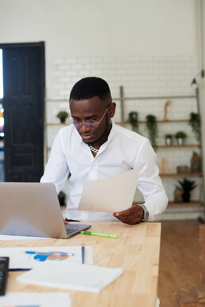 Nuori afrikkalainen johtaja paidassa istuu pöytänsä ääressä toimistossa, työskentelee verkossa kannettavan - Valokuva, kuva