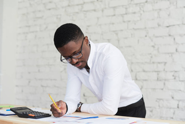 Gömlekli genç Afrikalı yönetici ofisinde masasında duruyor, kağıtlar ve grafiklerle çalışıyor. - Fotoğraf, Görsel