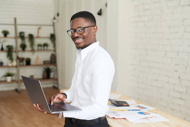 Lächelnder afrikanischer Mann, der in einem modernen Büro hinter einem Laptop arbeitet und in die Kamera blickt. Schöner afrikanisch-amerikanischer Geschäftsmann in Hemd und Hose nutzt Laptop und macht sich Notizen bei der Arbeit in - Foto, Bild