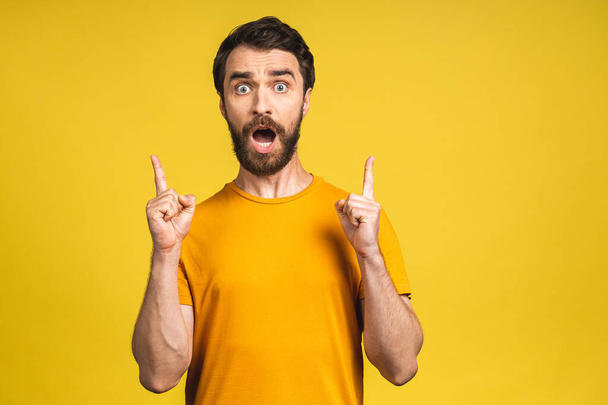 Look over there! Happy young bearded handsome man in casual pointing away up and smiling while standing isolated over yellow background. - Zdjęcie, obraz