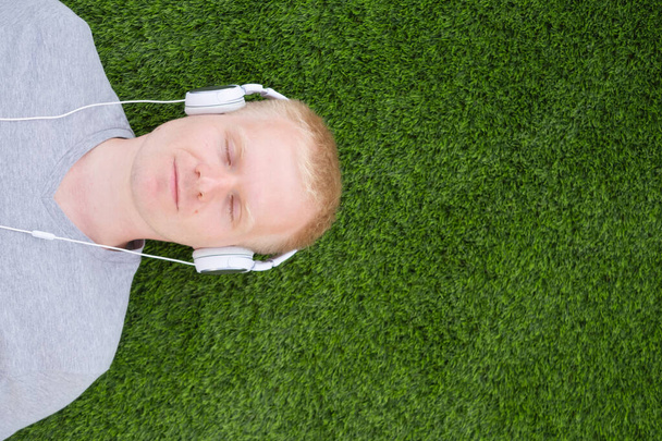 A young Caucasian man in a white T-shirt and headphones lies on an artificial lawn with closed eyes. The concept of listening to music, podcasts. Minimalism, copy space, top view.	 - Foto, Imagen