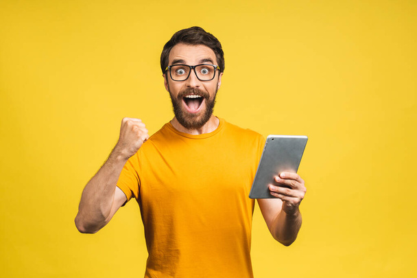 Happy young bearded man in casual standing and using tablet computer isolated over yellow background. - Foto, imagen