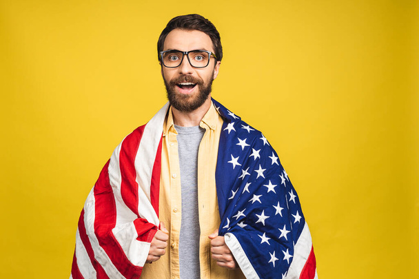 Portrait of young bearded happy man holding a USA flag isolated over yellow background. - Photo, image