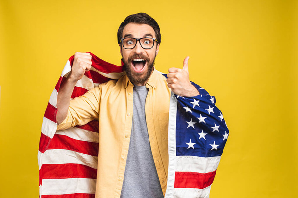 Portrait of young bearded happy man holding a USA american flag isolated over yellow background. - Fotografie, Obrázek