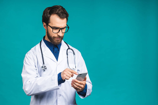 Молодой доктор пишет смс на смартфоне, изолированном на синем фоне. Использование мобильного телефона. - Фото, изображение