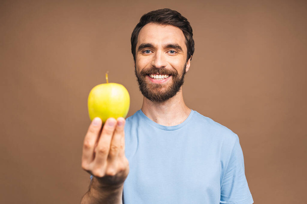 Έννοια διατροφής. Χαρούμενη όμορφη γενειοφόρος νεαρός άνδρας τρώει πράσινο μήλο, απομονώνονται σε μπεζ φόντο. - Φωτογραφία, εικόνα