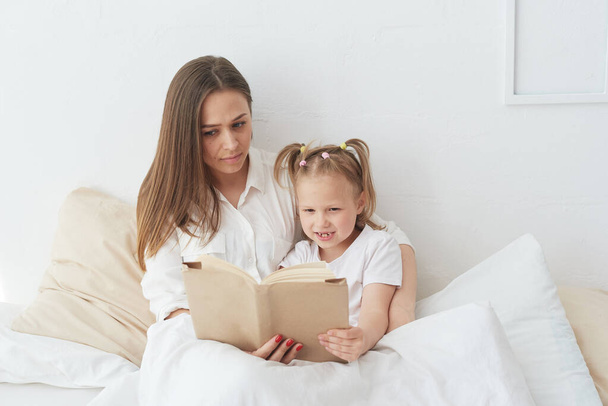 Young beautiful mother is reading a book to her daughter. Happy family, mother and daughter reading a book while lying in bed, smiling nanny mom telling funny story to cute preschool girl - Фото, изображение