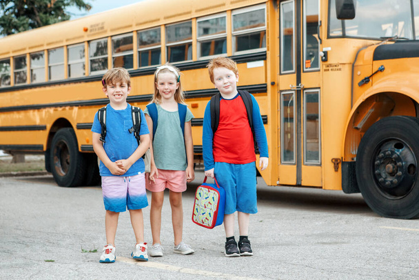 Onnellinen valkoihoinen lapset pojat ja tytöt lapset opiskelijat seisoo keltainen koulubussi. Koulutus ja takaisin kouluun syyskuussa. Ystävät luokkatoverit kokous kesäloman jälkeen. - Valokuva, kuva