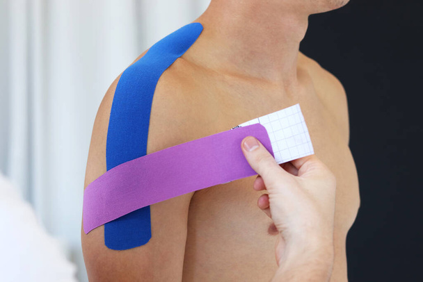 Kinesiology taping. Physical therapist applying kinesiology tape to patient shoulder. Male therapist treating injured shoulder of male athlete. Post traumatic rehabilitation, sport physical therapy. - Fotografie, Obrázek