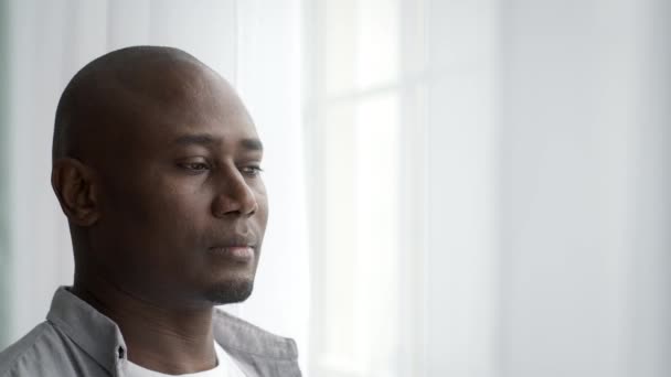 Pensativo hombre negro de mediana edad de pie cerca de la ventana en casa - Metraje, vídeo