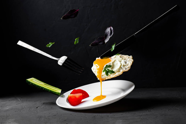 Fried egg levitation. Dark food photo. A chicken egg with liquid yolk, leaves of basil, dill, cucumber falls on a white plate. Breakfast creative concept. - Zdjęcie, obraz