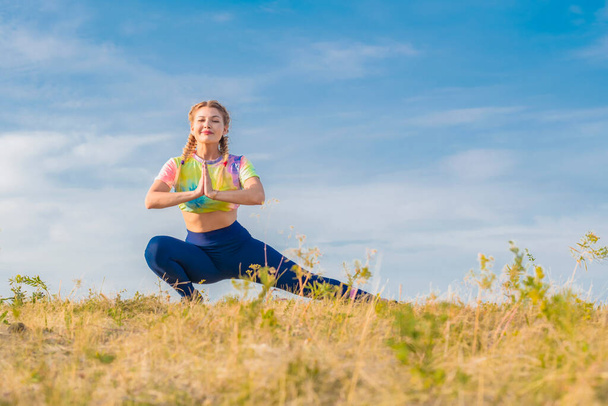 Young beautiful girl in multicolored tracksuit performs physical exercises to stretch muscles of legs outdoors on background of blue summer sky. Hands are folded in front of you. Looks into camera. - Valokuva, kuva