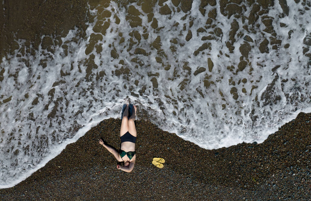 Woman with swimsuit resting on a sandy beach with braking waves on the shore. Overhead shot. Aerial drone photograph - Foto, afbeelding