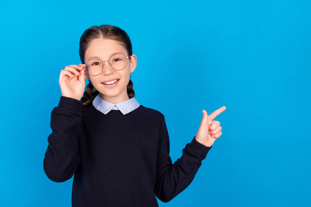 Photo of smiling good mood schoolgirl in eyeglasses point finger copyspace advertisement isolated on blue color background - 写真・画像