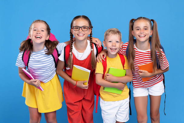 Πίσω στο σχολείο. Ευτυχισμένα παιδιά συμμαθητές, κορίτσια και αγόρια, στέκονται με σακίδια και βιβλία γυμναστικής μαζί και ποζάρουν για φωτογραφία, χαμογελώντας γενικά στην κάμερα, απομονωμένα σε μπλε φόντο στούντιο - Φωτογραφία, εικόνα