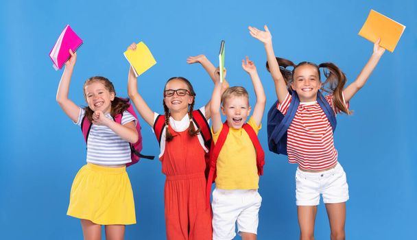 Lustige Klassenkameraden mit Rucksäcken springen am ersten Schultag vor Glück auf blauem Grund, heben die Hände mit Schulheften, Jungen und Mädchen freuen sich, nach den Ferien wieder in der Schule zu sein - Foto, Bild