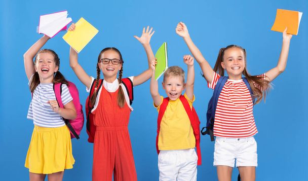 Funny kids classmates with backpacks jumping on blue background from happiness on first school day, raising hands up with exercise books, boys and girls excited to be back at school after vacation - Photo, image