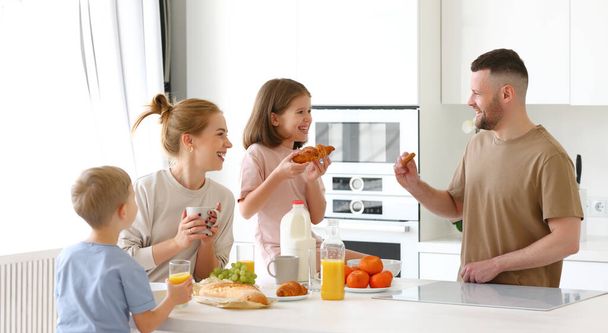 Young happy beautiful family having breakfast together at home. Father, mother and two cute little kids eating healthy food in morning, talking and smiling while standing in modern kitchen - Fotó, kép