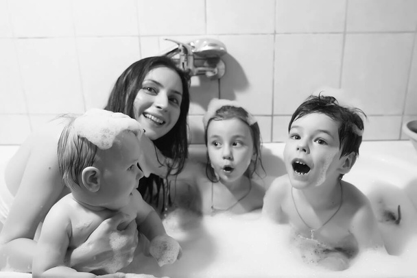 Η μαμά πλένει τα παιδιά. Η μαμά και τα παιδιά κάνουν επεμβάσεις νερού στο μπάνιο. Αφρός, σπρέι. - Φωτογραφία, εικόνα